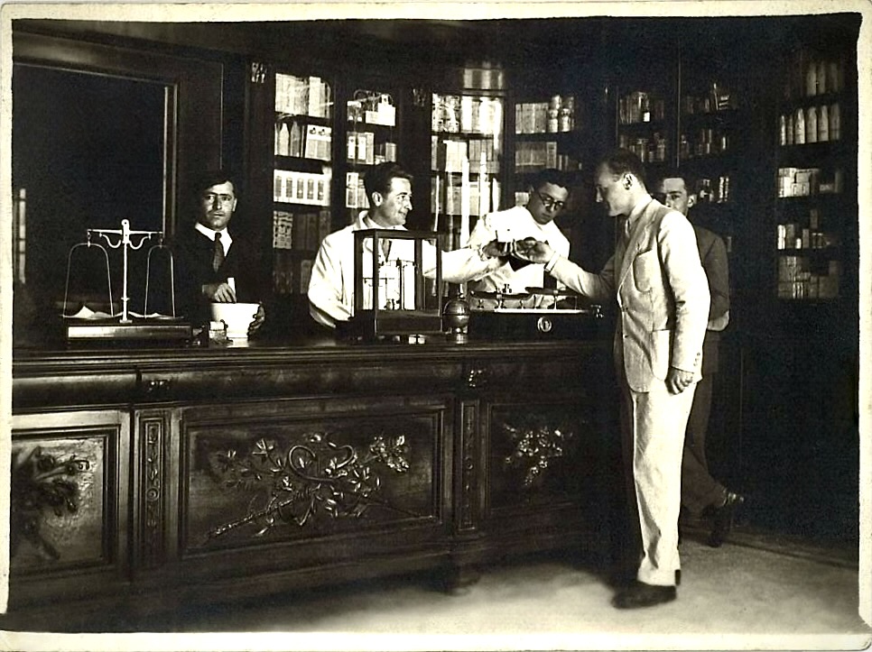 Il bancone della Farmacia Chiussi in una fotografia d'epoca, con il farmacista e un cliente al banco